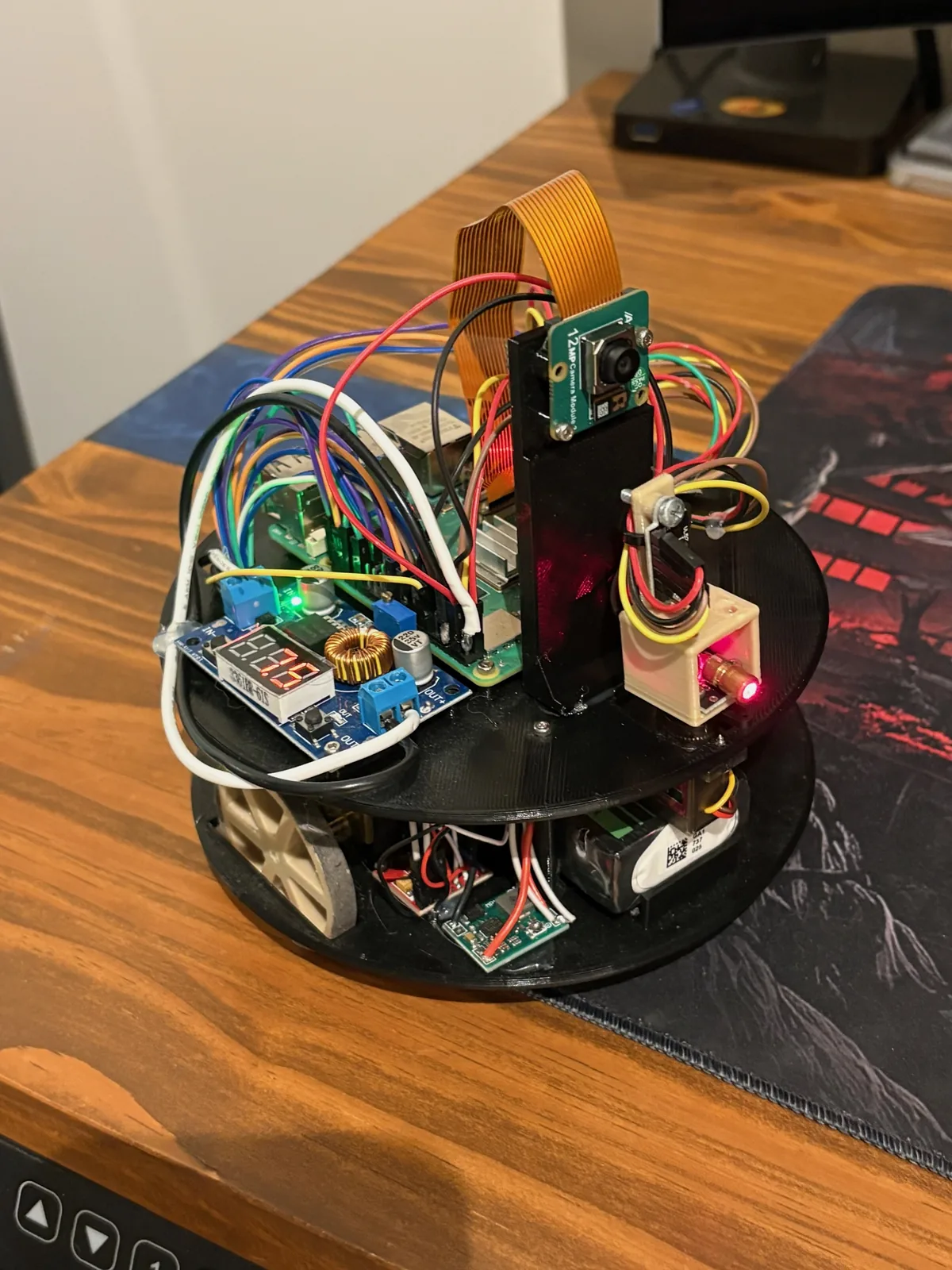 RoboTrail's top plate: a Raspberry Pi 5 with a camera module on a printed mast, a buck converter with a lit voltage display, the time-of-flight laser in a printed bracket, and the motor driver and battery on the deck below.